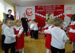 Na tle biało czerwonej dekoracji tańczą w parach dzieci ubrane na biało-czerwono.
